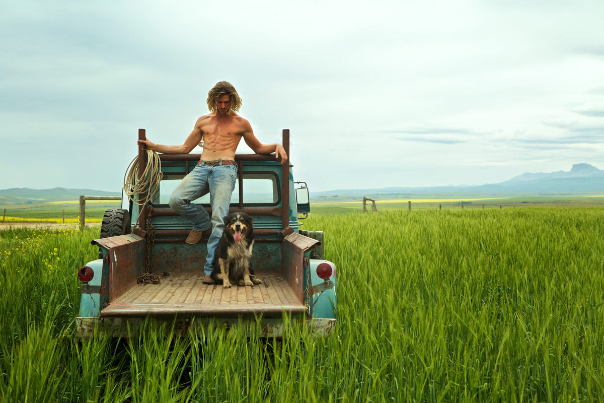 A man standing on the back of an old pickup in a prairie field, his dog on the tailgate